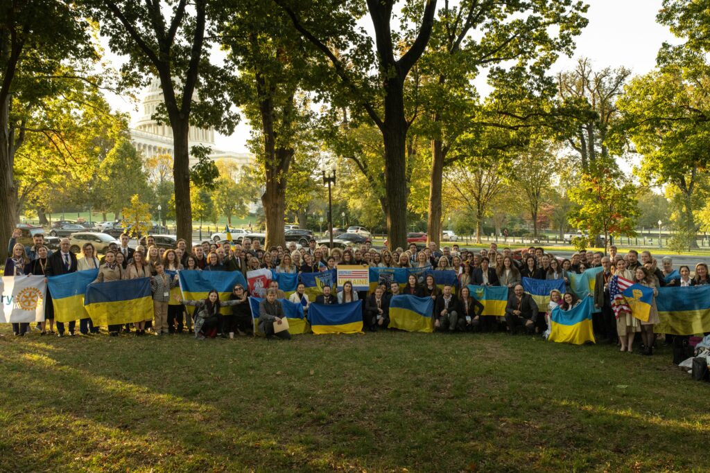 Lareg group of people standing in a line holding many Ukrainian flags with the US Capitol visible in the distance